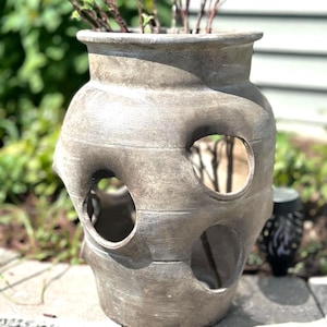 May include: A large, weathered gray ceramic planter with multiple circular openings. The planter has a wide, rounded body and a slightly flared rim. Green and brown plant stems emerge from the top. The planter is sitting on a stone surface.