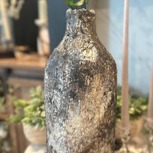 May include: A tall, cylindrical vase with a heavily textured, weathered surface featuring a mottled blend of dark charcoal, earthy brown, and off-white tones. The exterior has a rough, cracked appearance with a narrow neck and a wide opening.
