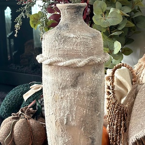 Puede incluir: Un jarrón cilíndrico alto con un acabado beige y gris muy texturizado y desgastado. El jarrón presenta un detalle horizontal prominente similar a una cuerda que rodea la sección superior, un cuello estrecho y una base ancha y robusta de piedra.