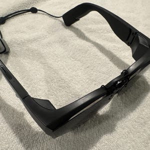 May include: Black smart glasses with a sleek, modern design. The glasses feature a dark frame, a nose bridge, and a connecting cord with a small control unit. The lenses are dark, and the overall aesthetic is minimalist and tech-forward.