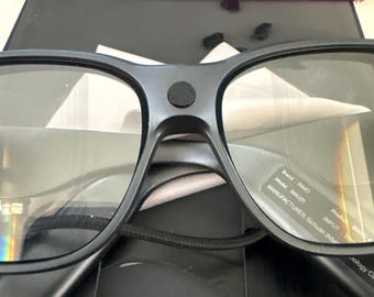 May include: Black smart glasses with a central circular sensor on the bridge, featuring a label on the lens with text including Brand INMO, Model IMA301, and Manufacturer Sichuan INMO. A purple precision tool lies in the background.