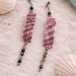 Può includere: Un paio di orecchini pendenti con pendenti rettangolari rosa strutturati e fili di perline colorate. Gli orecchini hanno borchie color argento e sono esposti sulla sabbia, con conchiglie. I pendenti sono in pietra.
