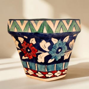 May include: A hand-painted ceramic flower pot featuring a wide rim with a green zigzag pattern. The body displays a dark blue background with red and blue floral motifs, white leaves, and a bottom border of red and white leaf shapes on teal.