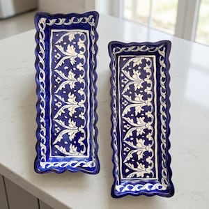 May include: Two rectangular ceramic serving trays featuring a deep blue glaze with white hand-painted floral and vine patterns. The trays have scalloped edges and a repeating wave-like border design, showcasing a glossy finish and elongated shape.