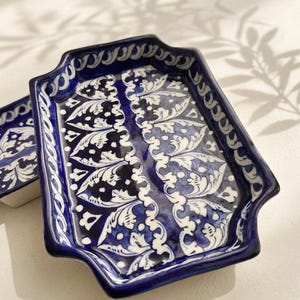Może przedstawiać: Prostokątna ceramiczna taca z falistym brzegiem, pokryta głęboką niebieską glazurą z misternymi, ręcznie malowanymi białymi wzorami kwiatów i liści. Wzór zawiera symetryczne zawijasy oraz motywy botaniczne na całej powierzchni.