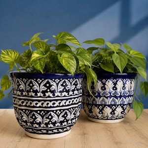 May include: Two round ceramic planters featuring a deep blue glaze with intricate white hand-painted floral and scroll patterns. Each pot contains a lush green pothos plant with variegated heart-shaped leaves spilling over the edges.