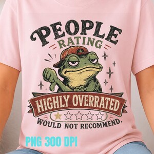Op de afbeelding: Lichtroze t-shirt met een afbeelding van een chagrijnige kikker met een pet en een duim omlaag. De tekst luidt "PEOPLE RATING HIGHLY OVERRATED WOULD NOT RECOMMEND". Het ontwerp bevat sterren en banners.
