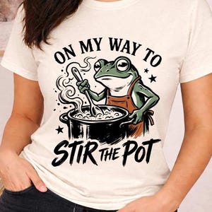 Könnte beinhalten: Cremefarbenes T-Shirt mit einer Cartoon-Fröschin, die eine Schürze trägt und in einem Topf rührt. Der Text "ON MY WAY TO STIR THE POT" ist oberhalb und unterhalb der Frosch-Illustration gedruckt. Das Design enthält Sterne und Dampf.