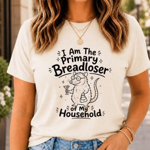 Pode incluir: Uma t-shirt de manga curta cor de creme com um gráfico preto de um gambá a usar óculos, segurando um copo de martini e uma mala. O texto diz: I Am The Primary Breadloser of My Household, rodeado por pequenos detalhes de estrelas.