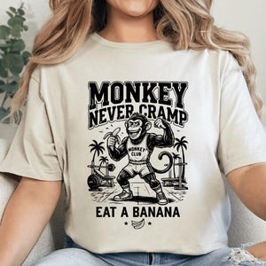 以下が含まれることがあります： クリーム色の半袖Tシャツで、ジムにいる筋肉質な猿の黒いグラフィックが描かれています。テキストは「MONKEY NEVER CRAMP EAT A BANANA」と書かれています。猿は「MONKEY CLUB」と書かれたタンクトップを着て、ヤシの木の近くでバナナを持っています。