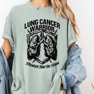 May include: A light green t-shirt with the words "LUNG CANCER WARRIOR" above a graphic of lungs and a shield. The text "STRONGER THAN THE STORM" is below the graphic. The design is in black.