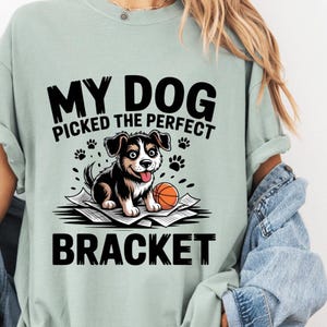 May include: A light green t-shirt with the text "MY DOG PICKED THE PERFECT BRACKET". The graphic features a cartoon dog with a basketball on top of papers. The dog is black and white with a happy expression.