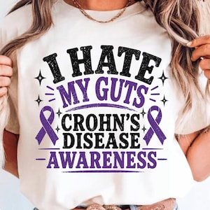 Op de afbeelding: Crèmekleurig T-shirt met de tekst "I HATE MY GUTS" in zwart en paars. Het shirt bevat de woorden "CROHN'S DISEASE AWARENESS" en twee paarse bewustzijns linten. Het ontwerp is gecentreerd.