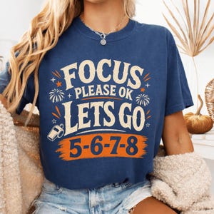 May include: A navy blue short-sleeve t-shirt featuring a graphic print in cream and orange. The text reads FOCUS PLEASE OK LETS GO 5-6-7-8. The design includes small star icons, firework illustrations, and a whistle icon on the left side.