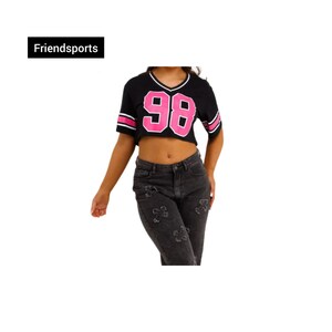 Può includere: Top corto nero con dettagli rosa e il numero 98. Il top ha uno scollo a V e maniche corte. Il testo "Friendsports" è in un rettangolo nero in alto a sinistra. La modella indossa jeans neri con disegni floreali.