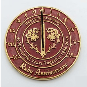 May include: A circular red and gold sundial featuring Roman numerals, a central gnomon, and engraved text reading Happy Anniversary, 40 Wonderful Years Together 1986-2026, and Ruby Anniversary, with a central floral design and two bells.