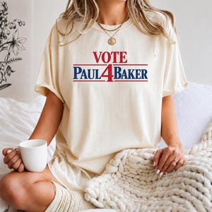 Könnte beinhalten: Ein cremefarbenes T-Shirt mit kurzen Ärmeln und dem in Rot und Blau auf die Brust gedruckten Text VOTE PAUL 4 BAKER. Das Design enthält horizontale Linien über und unter dem Namen, wobei die Zahl 4 den Buchstaben A im Namen ersetzt.
