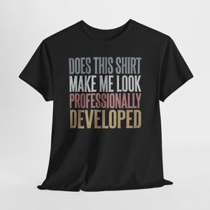 May include: A black short-sleeved t-shirt featuring the text "DOES THIS SHIRT MAKE ME LOOK PROFESSIONALLY DEVELOPED" printed in stacked lines. The words are coloured in light grey, white, muted pink, and gold, centred on the chest area.