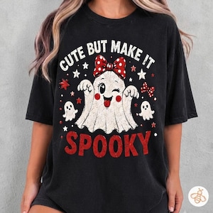 Puede incluir: Una camiseta negra de manga corta con el gráfico de un fantasma guiñando un ojo con un lazo rojo de lunares blancos, rodeado de fantasmas pequeños y estrellas. El texto "CUTE BUT MAKE IT" aparece arriba y "SPOOKY" está impreso abajo en rojo.