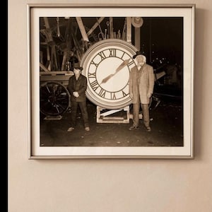 Può includere: Fotografia in bianco e nero incorniciata con due uomini in posa davanti a un grande orologio con numeri romani. Le lancette dell'orologio segnano le 10:04. Gli uomini indossano abiti d'epoca, uno con cappello e giacca a quadri.