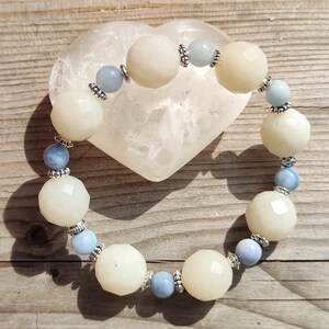 Könnte beinhalten: Ein Armband mit großen, facettierten, cremefarbenen Perlen, die mit kleineren, hellblauen Perlen abwechseln. Silberfarbene Abstandshalter trennen die Perlen. Das Armband wird auf einem herzförmigen, durchscheinenden Stein auf einer Holzoberfläche präsentiert.