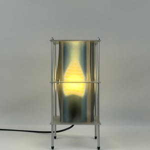 Peut inclure: Une lampe de table rectangulaire dotée d'un cadre en métal argenté avec quatre supports verticaux et deux étagères horizontales. Un panneau translucide incurvé avec un éclat dégradé bleu et jaune est placé entre les étagères, éclairé de l'intérieur.