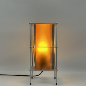 Puede incluir: Una lámpara de mesa rectangular con un marco de metal plateado que consta de cuatro soportes verticales y dos placas horizontales. Un panel translúcido y ondulado de color ámbar se sitúa en el interior, iluminado por una luz cálida interna.