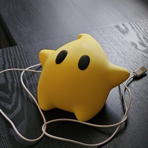 Op de afbeelding: Een gele ster-vormige nachtlamp met twee zwarte ogen en een USB-kabel. Het licht staat op een donker houten oppervlak. De ster heeft een afgeronde vorm en een kleine punt aan de bovenkant.