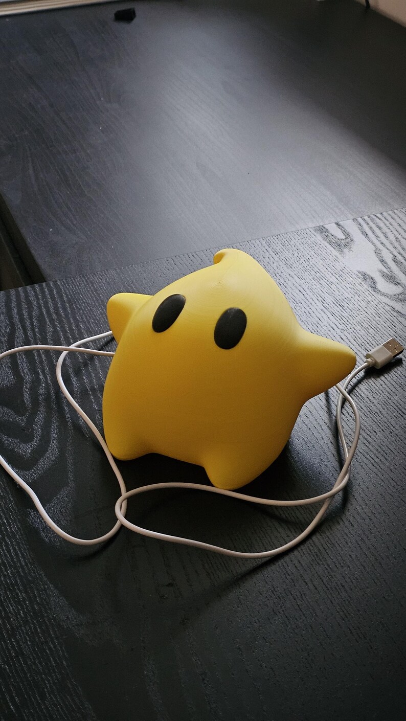 Op de afbeelding: Een gele ster-vormige nachtlamp met twee zwarte ogen en een USB-kabel. Het licht staat op een donker houten oppervlak. De ster heeft een afgeronde vorm en een kleine punt aan de bovenkant.