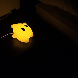 Op de afbeelding: Een geel nachtlampje in de vorm van een spook met twee zwarte ooguitsparingen. De lamp is aangesloten en straalt een zacht, warm licht uit tegen een donkere achtergrond. Het ontwerp is eenvoudig en kindvriendelijk.