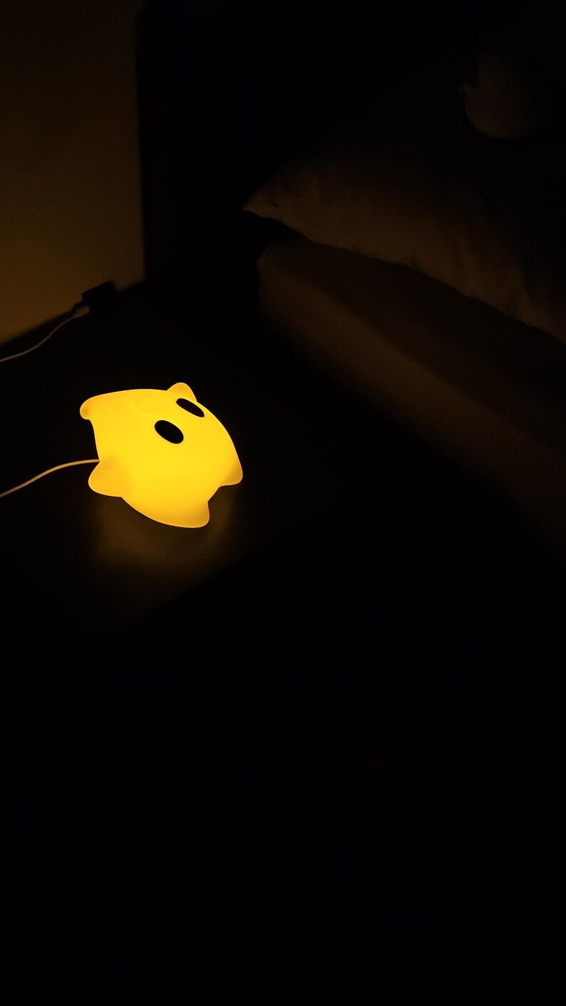 Op de afbeelding: Een geel nachtlampje in de vorm van een spook met twee zwarte ooguitsparingen. De lamp is aangesloten en straalt een zacht, warm licht uit tegen een donkere achtergrond. Het ontwerp is eenvoudig en kindvriendelijk.