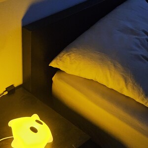 Op de afbeelding: Een gloeiend geel spookvormig nachtlampje staat op een donker nachtkastje naast een bed met een wit kussen en gele lakens. Het licht is aangesloten en geeft een warme gloed.