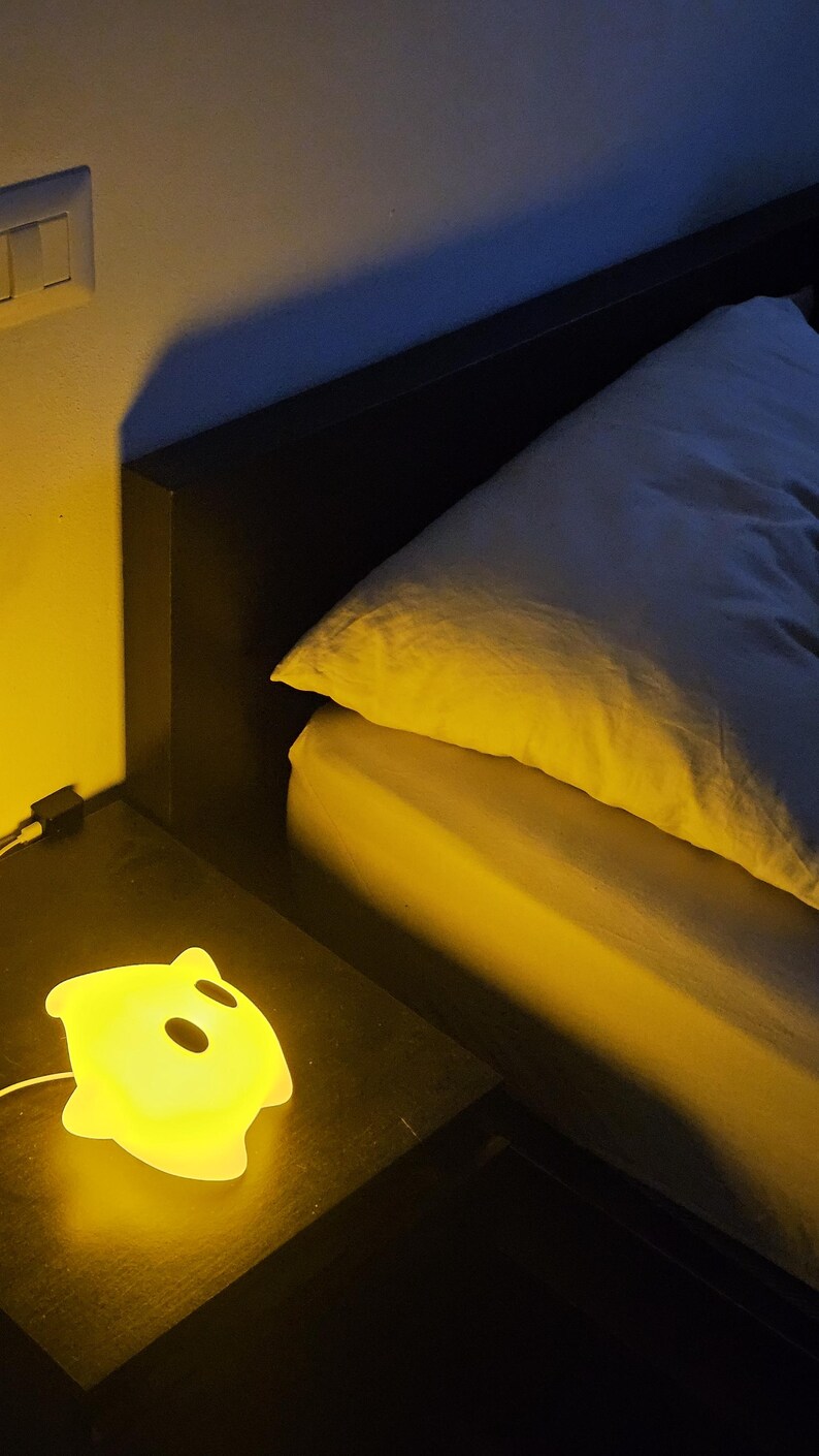 Op de afbeelding: Een gloeiend geel spookvormig nachtlampje staat op een donker nachtkastje naast een bed met een wit kussen en gele lakens. Het licht is aangesloten en geeft een warme gloed.
