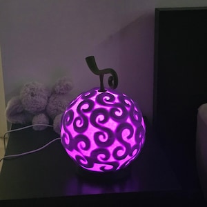 Può includere: Una lampada sferica viola brillante con un motivo a spirale, simile a un frutto stilizzato. La lampada ha uno stelo verde e poggia su una superficie scura. Un peluche è visibile sullo sfondo. La lampada è collegata.