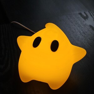 Op de afbeelding: Een gele ster-vormige nachtlamp met twee zwarte ovale ogen. De lamp geeft een warme gloed. De nachtlamp is gemaakt van een zacht materiaal en staat op een donker houten oppervlak.