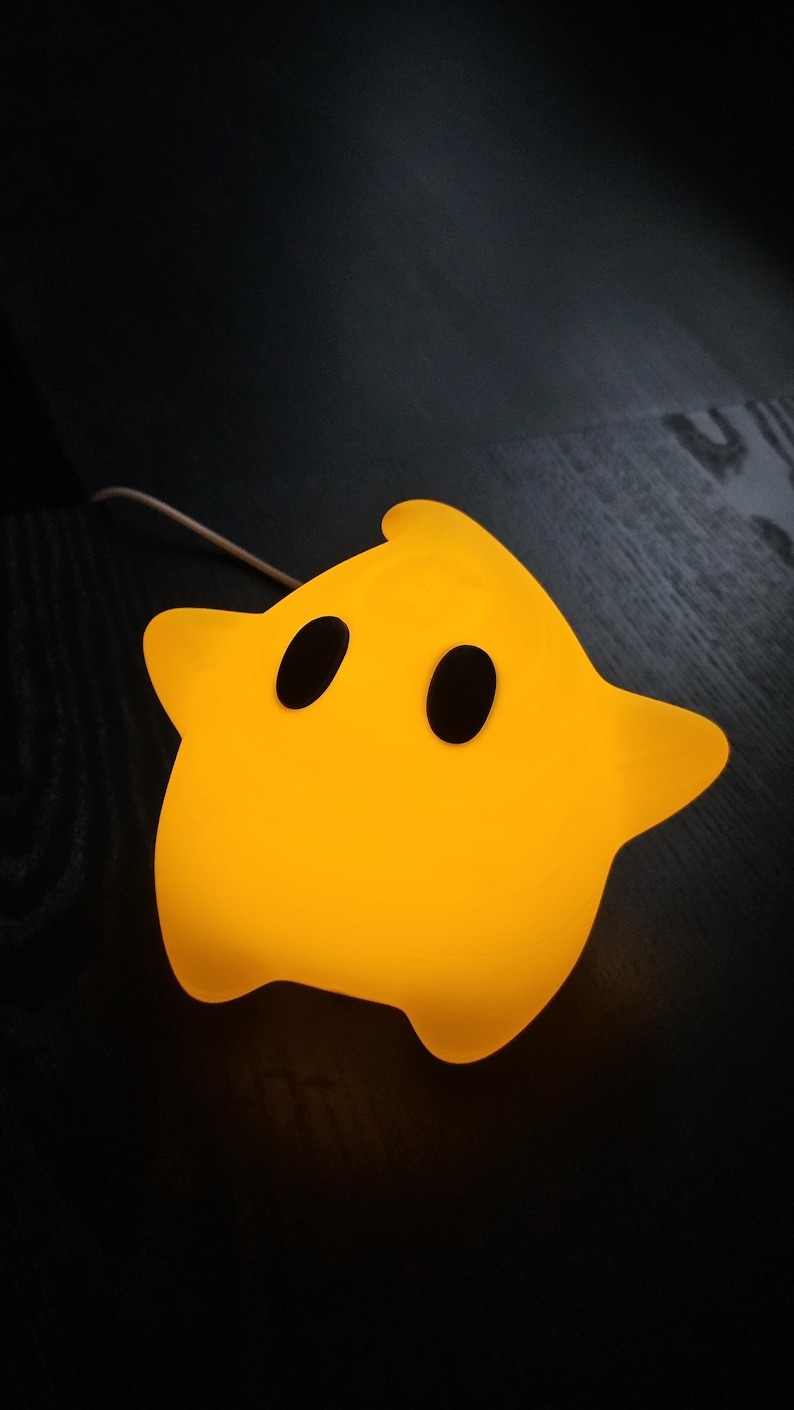 Op de afbeelding: Een gele ster-vormige nachtlamp met twee zwarte ovale ogen. De lamp geeft een warme gloed. De nachtlamp is gemaakt van een zacht materiaal en staat op een donker houten oppervlak.
