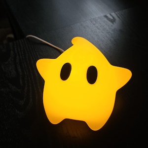 Op de afbeelding: Een gele ster-vormige nachtlamp met twee ovale zwarte ogen. Het licht gloeit en heeft een afgeronde vorm. De nachtlamp staat op een donkere ondergrond en heeft een wit snoer.