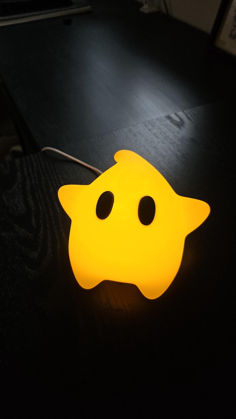 Op de afbeelding: Een gele ster-vormige nachtlamp met twee ovale zwarte ogen. Het licht gloeit en heeft een afgeronde vorm. De nachtlamp staat op een donkere ondergrond en heeft een wit snoer.