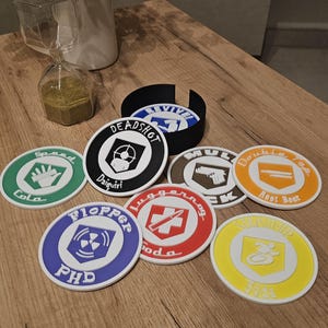 Può includere: Un set di sottobicchieri rotondi con vari disegni e testi, tra cui "Deadshot Daiquiri" e "Speed Cola". I sottobicchieri sono in un supporto, su una superficie di legno.