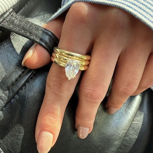 Op de afbeelding: Een hand met drie gouden ringen, waarvan één met een peervormige diamant. De ringen zijn op de ringvinger gestapeld. De achtergrond omvat een zwart leren oppervlak en een blauw-wit gestreept overhemd.