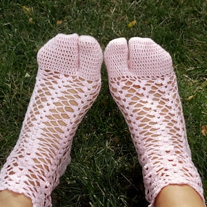 May include: A pair of light pink crochet socks featuring a split-toe design. The socks are crafted with a delicate, open-work lattice pattern that extends from the ankle down to the foot, ending in a solid, closed-toe section for the big toe.
