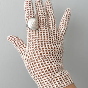 May include: A cream-coloured, open-weave crochet glove with a silver ring featuring a white, spiral seashell. The glove is made of a delicate, net-like material. The ring is worn on the middle finger.