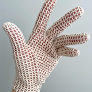 K&ouml;nnte beinhalten: Ein beigefarbener H&auml;kelhandschuh mit offenem, gitterartigem Gewebe. Der Handschuh besteht aus einem hellfarbigen Garn. Die Finger sind vollst&auml;ndig geformt, und der Handschuh scheint an einer Hand getragen zu werden.