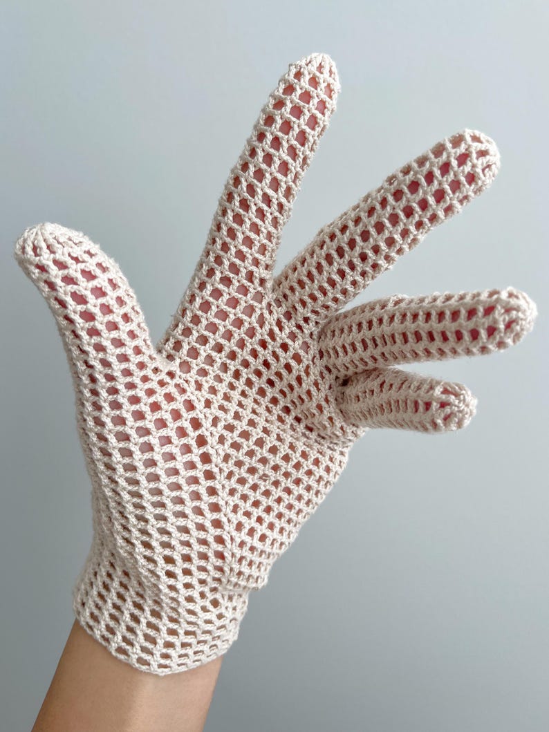 K&ouml;nnte beinhalten: Ein beigefarbener H&auml;kelhandschuh mit offenem, gitterartigem Gewebe. Der Handschuh besteht aus einem hellfarbigen Garn. Die Finger sind vollst&auml;ndig geformt, und der Handschuh scheint an einer Hand getragen zu werden.
