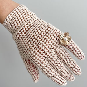 K&ouml;nnte beinhalten: Cremefarbene, offen gewebte H&auml;kelhandschuhe mit einer goldfarbenen Blumenbrosche mit Perlenmittelpunkt. Die Handschuhe haben ein zartes, netzartiges Muster und bedecken Hand und Handgelenk. Die Brosche ist auf der R&uuml;ckseite der Hand am Handschuh befestigt.