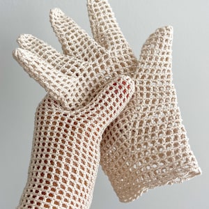 K&ouml;nnte beinhalten: Ein Paar cremefarbene, offen gewebte H&auml;kelhandschuhe. Die Handschuhe haben ein zartes, netzartiges Muster mit einem gewellten Rand am Handgelenk. Die Finger sind vollst&auml;ndig geformt und die Handschuhe scheinen handgefertigt zu sein. Der Hintergrund ist hell.