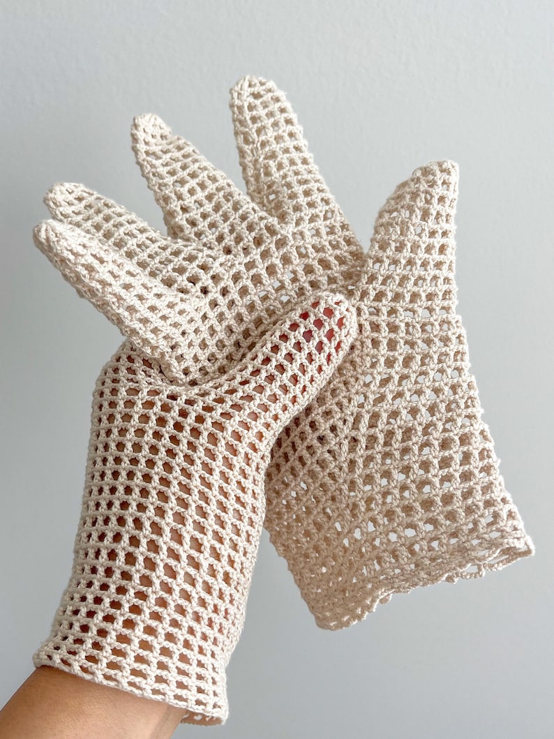 K&ouml;nnte beinhalten: Ein Paar cremefarbene, offen gewebte H&auml;kelhandschuhe. Die Handschuhe haben ein zartes, netzartiges Muster mit einem gewellten Rand am Handgelenk. Die Finger sind vollst&auml;ndig geformt und die Handschuhe scheinen handgefertigt zu sein. Der Hintergrund ist hell.
