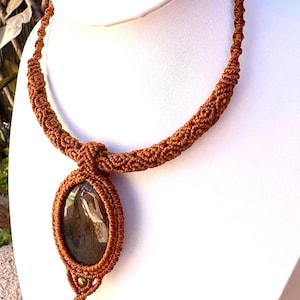 Puede incluir: Collar de macramé marrón hecho a mano con un colgante ovalado y un cordón tejido. El colgante contiene una piedra oscura con patrones, y un dije en espiral cuelga debajo. El collar se exhibe en un busto blanco.