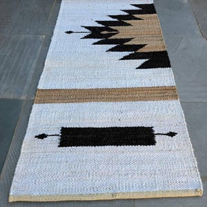 Op de afbeelding: Een opgerolde, rechthoekige witte loper met een geometrisch ontwerp. De loper heeft een zwart en bruin patroon in het midden, met een zwarte rechthoek aan elk uiteinde. De loper is gemaakt van geweven materiaal en wordt gepresenteerd op een grijze ondergrond.