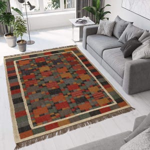 May include: Rectangular area rug with a geometric pattern in shades of red, orange, green, gray, and black. The rug has a cream-colored border and fringe along the edges. The rug is placed on a white wooden floor.